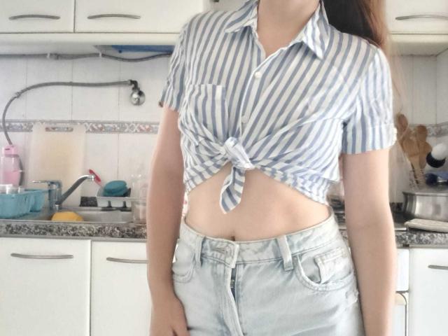 BraniaRuben with a blue and white striped shirt tied just below her tits, showing her midriff, wearing light denim shorts, standing in a kitchen.