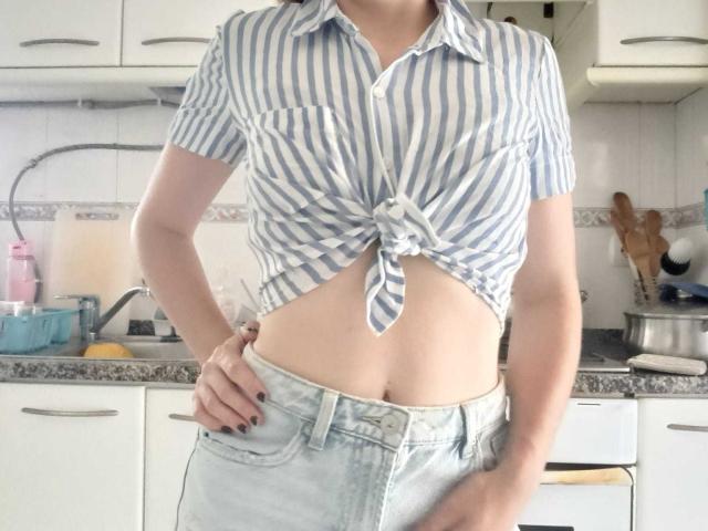 BraniaRuben with tied blue-striped shirt exposing her flat midriff, wearing light denim jeans, standing in a kitchen, hand on hip, no visible tits or pussy.