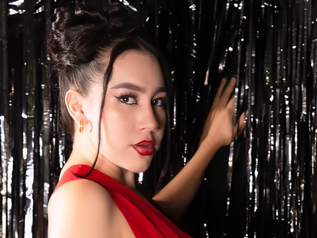 CelesteRusso with dark hair in a bun, red lipstick, and a red dress, touching black shiny curtain with her right hand, looking over her shoulder.