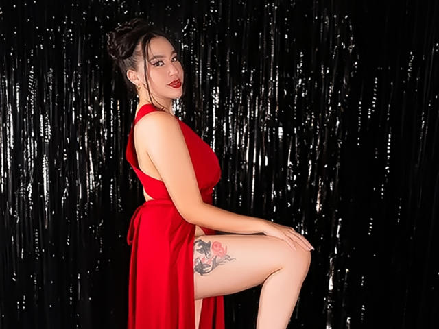 CelesteRusso in a red dress, showing her tits and pussy tattoo, sitting with one leg up, black curtain background, red lipstick, and dark hair in a bun.