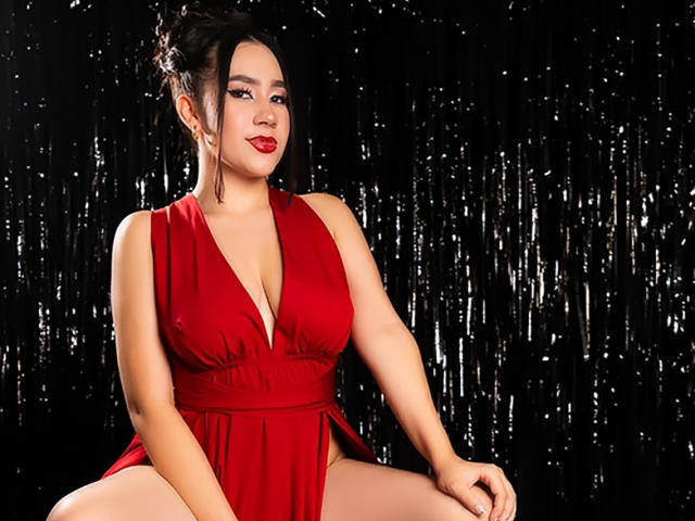 CelesteRusso with large boobs, wearing a red dress, sitting with legs apart, black shiny background, dark hair in a bun, red lipstick, looking at camera, confident smile.