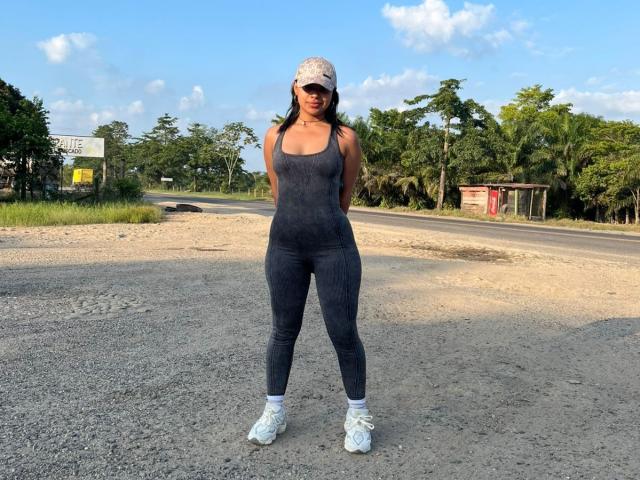SoleneFontaine with dark gray, tight, sleeveless, and legging outfit, white sneakers, and a pink cap, standing on gravel with hands behind her back, wearing sunglasses.