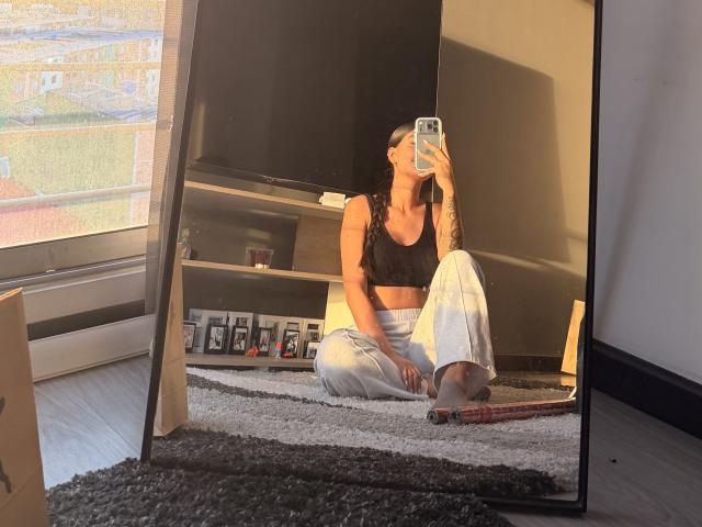 ThaliaAres mit langem, schwarzem Haar, die einen schwarzen Sport-BH und weiße Hosen trägt, sitzt im Schneidersitz und macht ein Selfie mit ihrem Handy, wobei ihre Tattoos und ihre kleinen Brüste zu sehen sind..