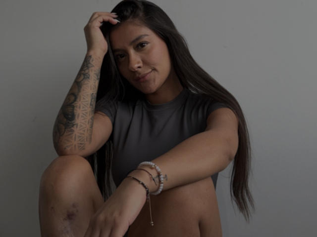 ThaliaAres with long dark hair, tattooed arm, grey t-shirt, and bracelets, sitting with knee up, hand in hair, slight smile, grey background. Boobs not visible, pussy not visible.