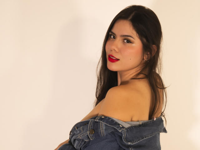 MaraCortes with long dark hair, red lipstick, and a denim jacket off her shoulders, looking over her shoulder with a subtle smile, showing her bare shoulders and upper chest.