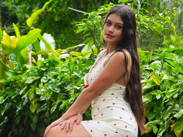 LizzaRose with long dark hair, white dress, small red flower pattern, sitting on a green leafy bush, looking over her shoulder, cute, pretty, soft smile, slightly curvy.