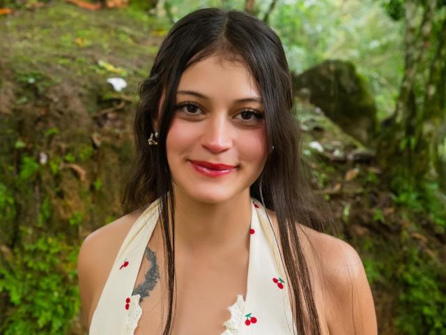 LizzaRose has long dark hair, red lipstick, and a small smile. She's wearing a white halter top with red cherries. Her tits are partially visible. She has a tattoo.