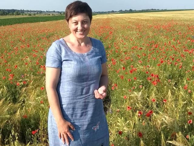 EmilyLlove, capelli castani corti, sorridente, indossa un vestito blu, in piedi in un campo di papaveri rossi, erba verde e grano, tiene un fiore bianco, giornata di sole, cielo blu e alberi in lontananza..