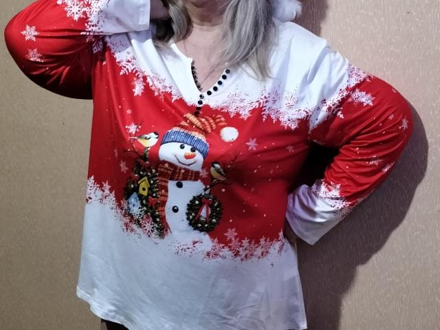 LolkaAlyss wears a red and white holiday shirt with snowflake patterns. The shirt features a cute snowman graphic with a blue hat and red scarf, surrounded by a green wreath. The red upper section transitions to white at the bottom. LolkaAlyss has blonde hair and stands with one hand on her hip, partially visible. The shirt's texture is slightly shiny. The background is a plain, beige wall.