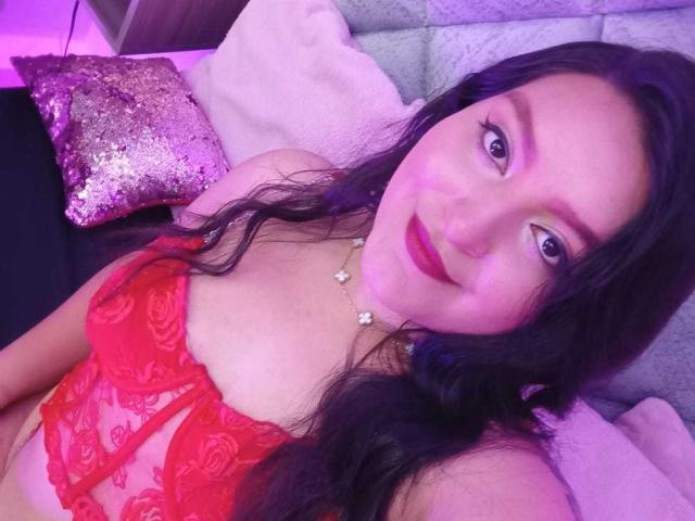 VictoriaRiver with long black hair, red lace bra, and red lipstick, smiling at the camera, lying on a bed with a sequined pillow, wearing a necklace with small flowers.