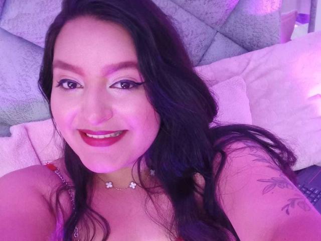 VictoriaRiver with long black hair, purple-pink lighting, smiling, dark red lipstick, wearing a necklace, tattoo on right arm, visible cleavage, sitting on a bed, smiling at camera.