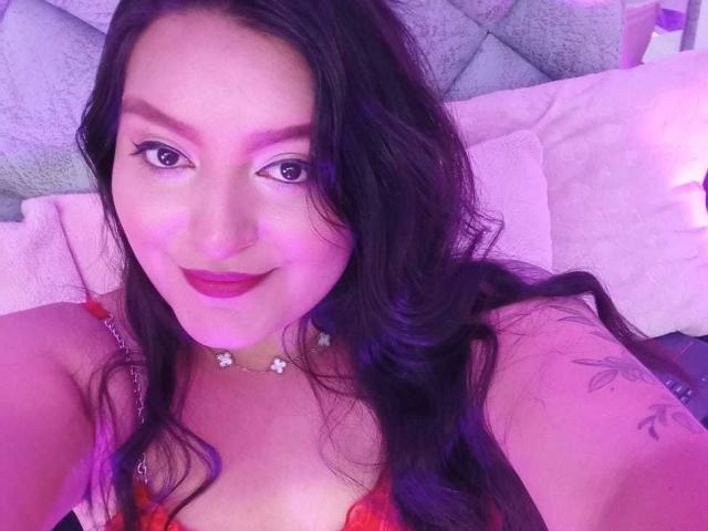 VictoriaRiver with purple-tinted skin, long dark hair, red lipstick, floral necklace, and tattoo on right arm, smiling at camera, wearing red lace bra, purple lighting.