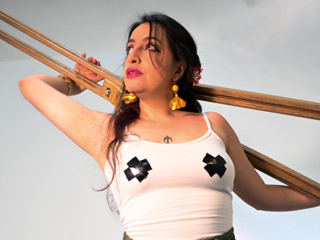 RozarioPinceles with dark hair, pink lipstick, white tank top, black nipple pasties, yellow earrings, holding wooden poles, looking up, tattoo on chest, medium boobs, medium skin, daytime.