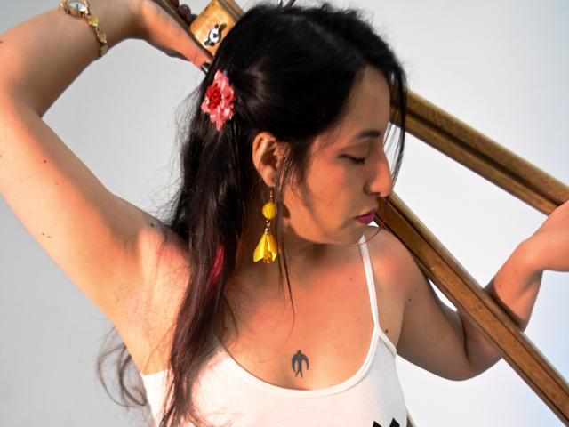 RozarioPinceles with black hair, white tank top, yellow tassel earrings, pink flower in hair, holding wooden stick, arm raised, small tattoo on chest, bracelet on wrist, eyes closed.