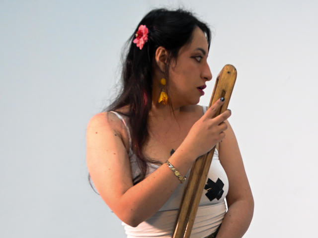 RozarioPinceles holds a wooden crossbow-like object with her right hand. She has long black hair with a pink flower and wears yellow triangular earrings. She's dressed in a white tank top with black cross pasties. Her right wrist has a gold bracelet. Her skin is light, and she looks to the right. The image is a photograph with a clear, light blue background.