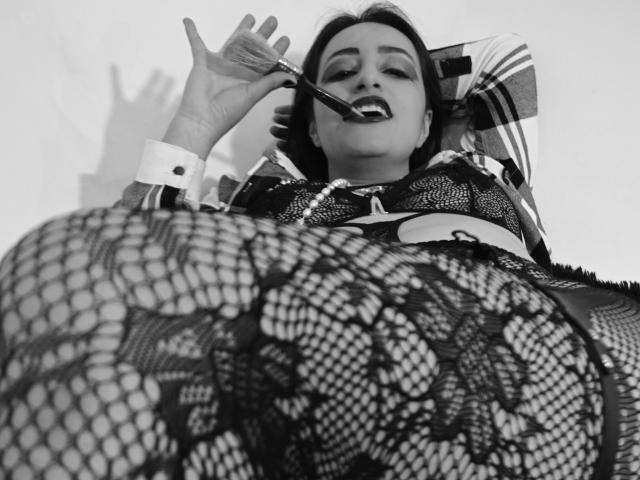 RozarioPinceles with a cigarette in her mouth, wearing a lace bra and fishnet stockings, lying back with her ass in the foreground, looking up with a relaxed expression.