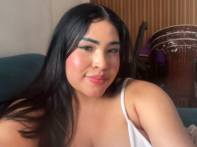 OliviGrace has dark, straight hair, a pink blush on her cheeks, and is smiling with her pink lips. She's wearing a white tank top, showing her small tits.