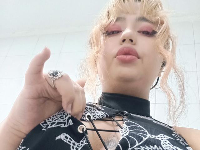 BelleVivienneHt heeft blond haar, roze oogschaduw en volle lippen. Ze draagt een zwart top met een patroon en een veter sluiting, en heeft een ring aan haar vinger..