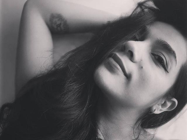 BronaaSexy has long, dark hair, a slight smile, and a tattoo on her arm. She's looking up with her arm behind her head in a black-and-white photo.