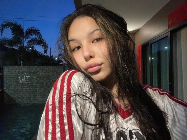 Close-up of MissSinful, a young Asian woman with long, wet, dark brown hair, standing near a pool at dusk. She has a neutral expression, slightly pursed lips, and is wearing a white and red striped sports jersey. Her skin is light, and her eyebrows are thin. The background includes a stone wall, palm trees, and a red wall section. The image has a natural, slightly overexposed light source from the right.