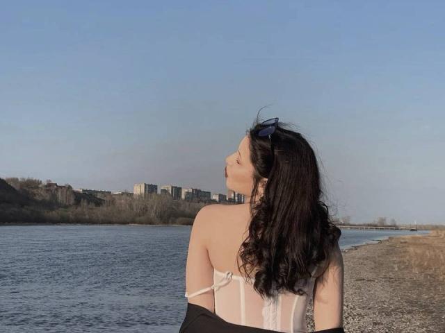 MissSinful with wavy black hair, wearing a white strapless top, stands by a river, looking up, with blue sunglasses on her head, and a black jacket around her elbows.