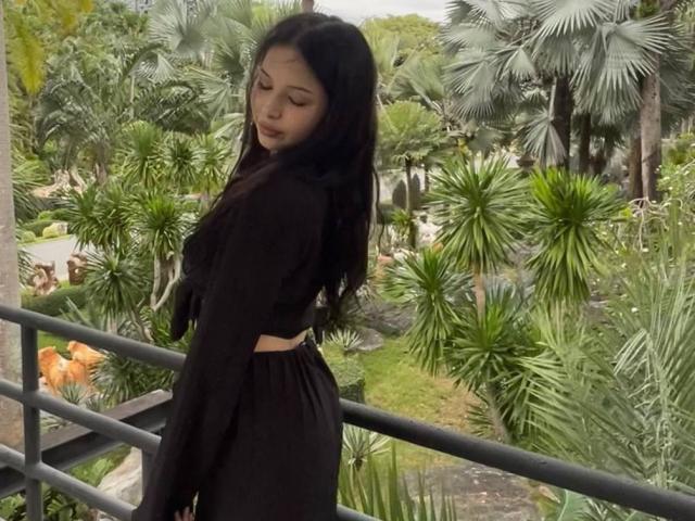 MissSinful with long black hair, wearing a black long-sleeve top and black pants, standing on a balcony, looking down, surrounded by green palm trees and foliage.