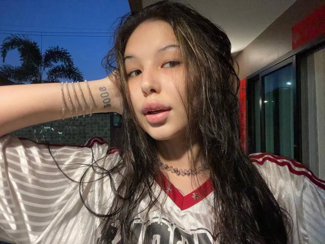 MissSinful, avec ses cheveux noirs mouillés, portant un maillot blanc brillant, a un tatouage sur le bras, un collier et des bracelets, et regarde l'appareil photo avec la bouche légèrement ouverte..