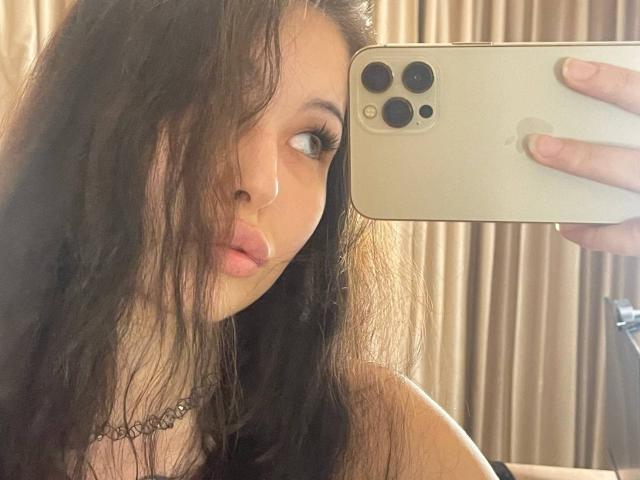 MissSinful with long, wavy dark hair, pouty pink lips, and a black choker, taking a mirror selfie with a gold iPhone, showing her face and upper chest.