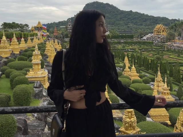 A photograph of MissSinful standing in a garden with golden stupa-like structures. She has long, dark hair, wearing a black, long-sleeved top with a midriff and black pants. Her right arm rests on a black railing. The background features green, manicured hedges, golden statues, and a mountain. The sky is overcast. MissSinful is looking to the left. The image uses natural light, emphasizing the contrast between her dark clothing and the golden structures. Her right hand is partially hidden under her left arm.