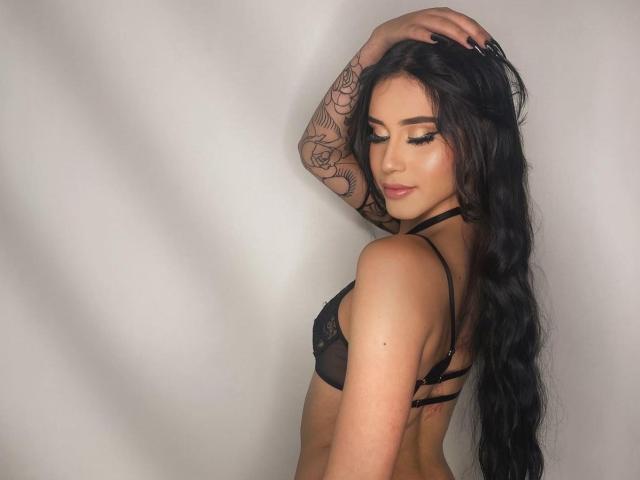 EmilyyRousee with long black hair, wearing a sheer black bra, exposing her tits, tattooed arm, and sexy makeup, standing with her back to the camera, eyes closed.