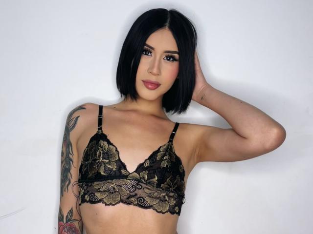 EmilyyRousee with black bob, wearing lace bra, hand in hair, tattooed arm, looking at camera, small tits, pale skin, subtle makeup, pink lipstick, white background.