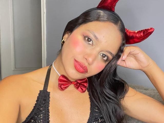 JeimyCampos with red devil horns, black lace top, red bowtie, red lipstick, and blushed cheeks, posing with one hand on her head, showing off her tits.