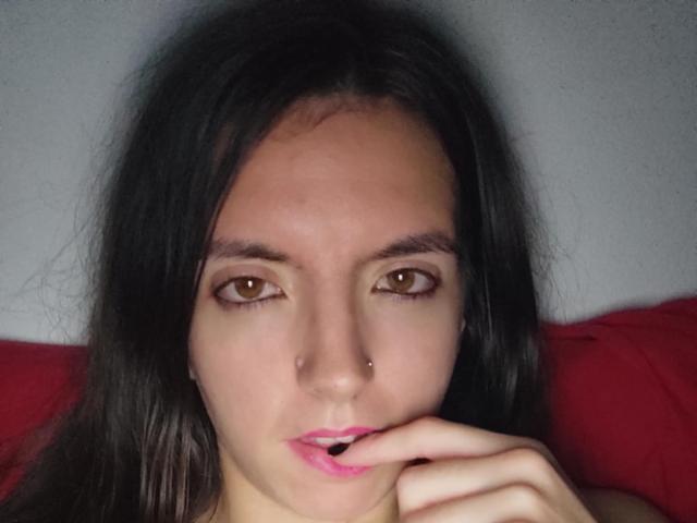 BraniaRuben with brown eyes, black hair, nose piercing, and pink lips, gently biting her pink-manicured finger. She's looking straight at the camera with a slight pout.