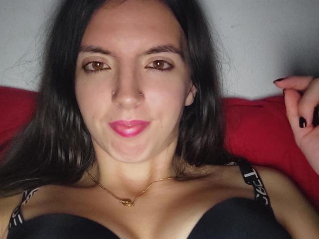 BraniaRuben with brown eyes, black hair, nose ring, pink lipstick, black bra, gold necklace, lying on red pillow, black nails, slightly smiling, upper body, chest visible.