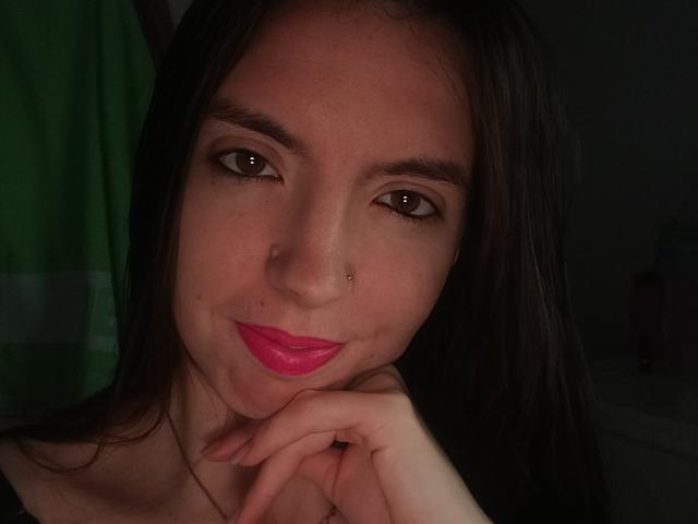 BraniaRuben with dark brown eyes, pink lipstick, nose piercing, and long brown hair, resting chin on hand, smiling softly in dim light, showing subtle makeup and relaxed expression.