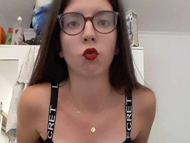 BraniaRuben with red lipstick, pouty lips, glasses, and black CREET bra straps, wearing a gold necklace, has long brown hair, fair skin, and a cute face.