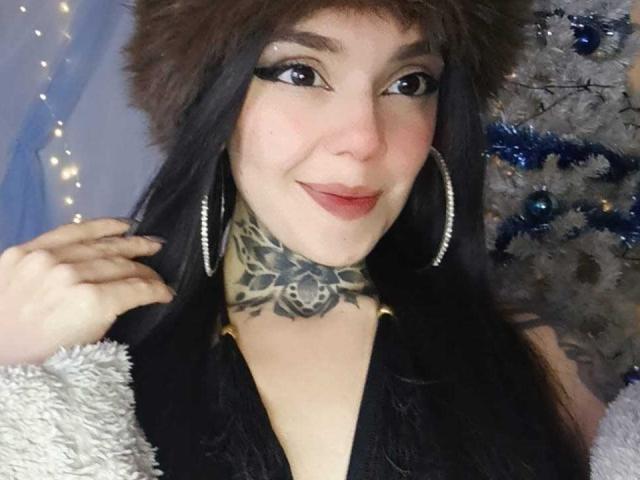 Lannie Gonzales, com cabelo escuro, chapéu de pele, delineador preto, brincos de argola, pescoço tatuado, sorrindo, batom vermelho, blusa preta, manga branca felpuda, mão direita tocando o chapéu, árvore de Natal ao fundo..