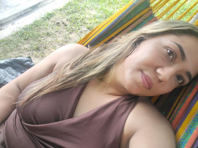 ViolaBertie with blonde hair, brown eyes, and a nose ring, wearing a brown sleeveless top, lying on a colorful striped hammock, looking at the camera with a slight smile.