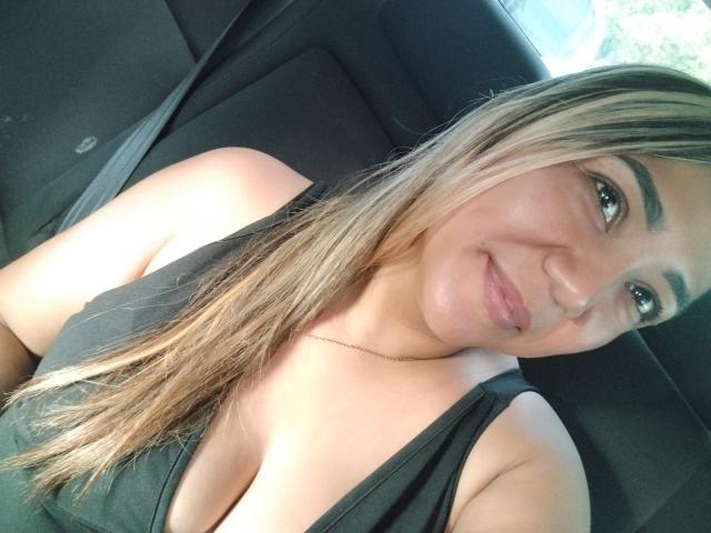 ViolaBertie with blonde hair, black tank top, and visible cleavage, smiling in a car seat, wearing a thin necklace, looking at the camera.