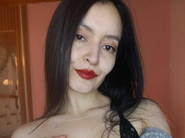 AriaBonnet with long black hair, red lipstick, and a slight smile, wearing a black lace top, has her tits barely covered by a gray sleeve.