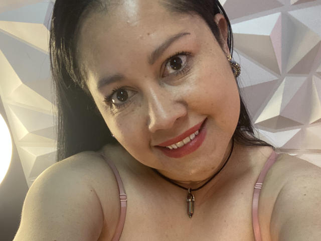 Close-up of MarianaSmitt with light skin and straight black hair, smiling at the camera. She has brown eyes, thin eyebrows, and red lipstick. MarianaSmitt wears a pink spaghetti strap top and a black choker with a small pendant. Geometric patterned background is visible. Her skin is slightly shiny, and she has small freckles on her nose. Her ears have small, ornate earrings. The image is a selfie, with a slight upward angle.