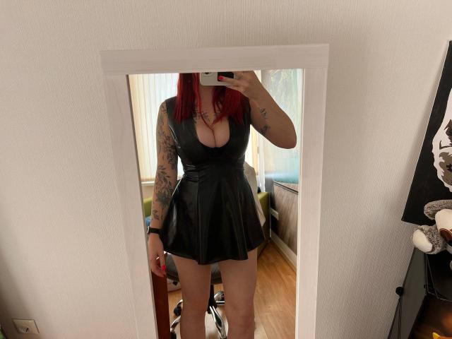 Innysik with red hair, tattoos on arms, wearing black, shiny, cleavage-revealing dress, mirror selfie, holding phone, visible thighs, wooden floor, white wall, partial chair in background.