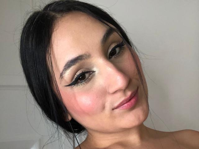HiilaryDolce has black hair, brown eyes, bold eyeliner, shiny eyeshadow, pink cheeks, and a slight smile, with bare shoulders, looking at the camera.