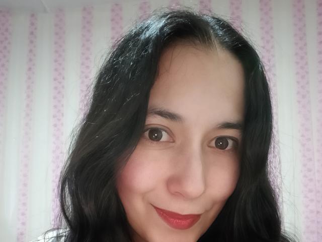 AnneStone has dark, wavy hair, brown eyes, and a subtle smile with red lipstick. Her skin is light with a slight blush on her cheeks. She's looking at the camera.