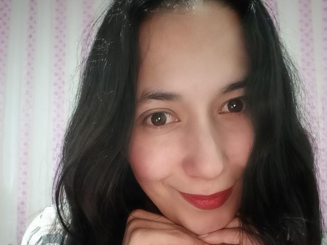 AnneStone with black wavy hair, red lipstick, and a cute smile, resting her chin on her hands, showing her soft pink cheeks and dark brown eyes.