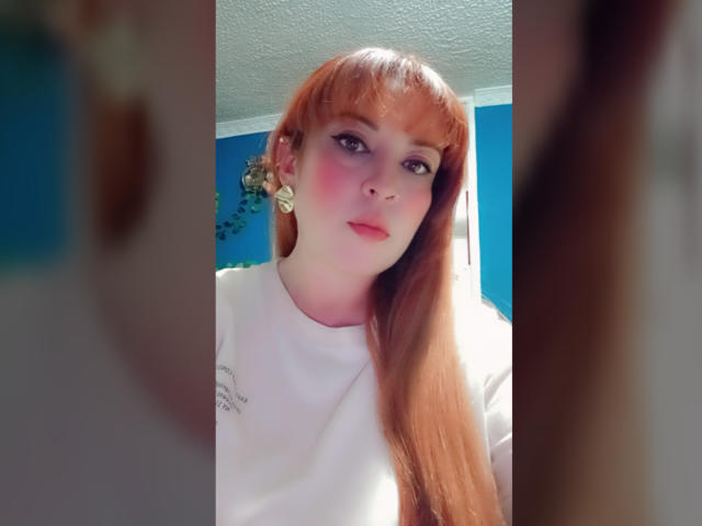 RosseHot69 con cabello rojo largo, rubor rosado en las mejillas, camisa blanca y aretes dorados, mirando a la cámara con una ligera sonrisa, pared azul de fondo..
