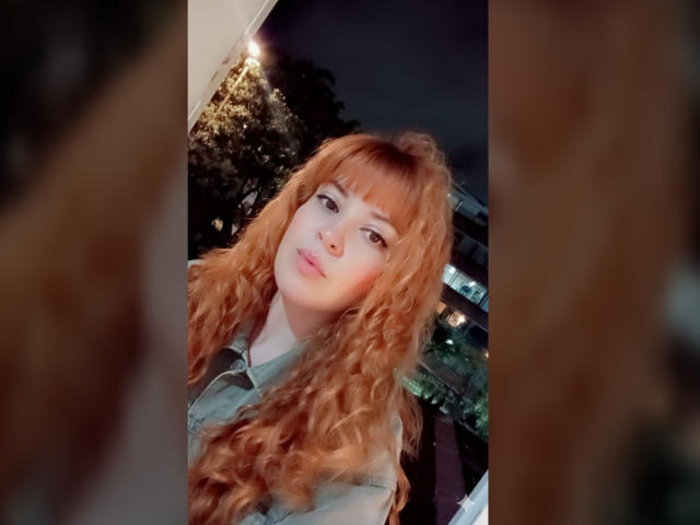 RosseHot69, avec de longs cheveux roux bouclés, une peau pâle et des yeux verts, portant une chemise verte, avec une expression mignonne et légèrement satisfaite, dans une scène nocturne peu éclairée..