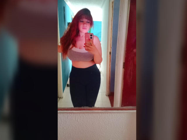 RosseHot69, com cabelo ruivo, vestindo um top esportivo cinza e calças pretas, em frente a um espelho, segurando um celular, mostrando seus seios e nádegas..