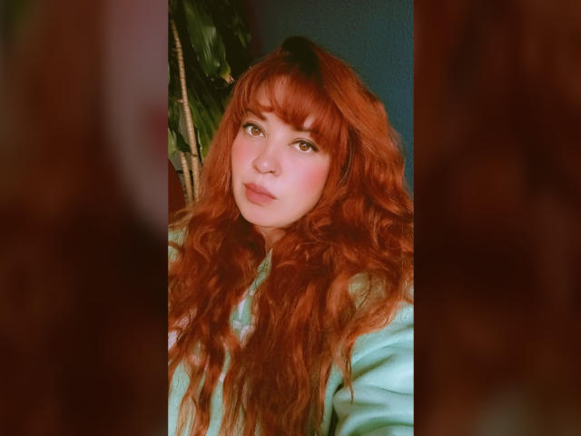 A portrait of RosseHot6. She has long, wavy red hair with blunt bangs, and fair skin. Her green eyes look slightly to the right. She wears a light green top. The background is blurred, with a dark green leafy plant on the left. The image has a soft, warm filter, enhancing the natural tones of her hair and skin. The focus is on RosseHot6's face, with a slight tilt to the right. The overall composition is balanced and centered.