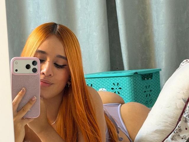 EmmaWat takes a selfie with a pink phone case in a mirror. She has long orange hair, closed eyes, and pink lipstick. She's lying on a bed, wearing a purple tank top and purple panties, with her butt slightly raised. A turquoise laundry basket with a floral pattern is in the background. Her right hand holds the phone, and her left arm is partially visible. The image has a casual, intimate feel.